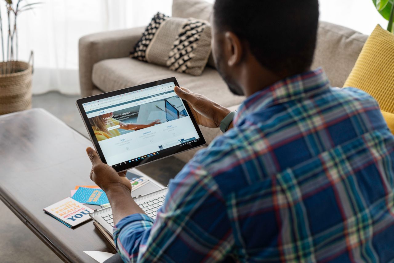 Man with plaid shirt browsing internet on tablet at home, seated on sofa.